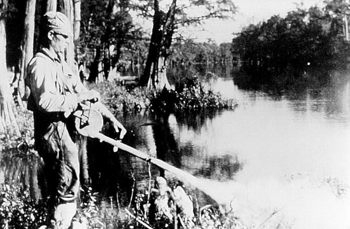 A man in a cap and work clothes stands at the edge of a wooded bayou and sprays a mist from a hand-held applicator out over the water.